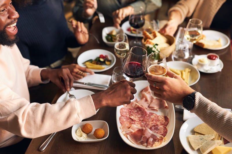 People toasting at a dining table with food and drinks. Black man smiles, holding up a glass.