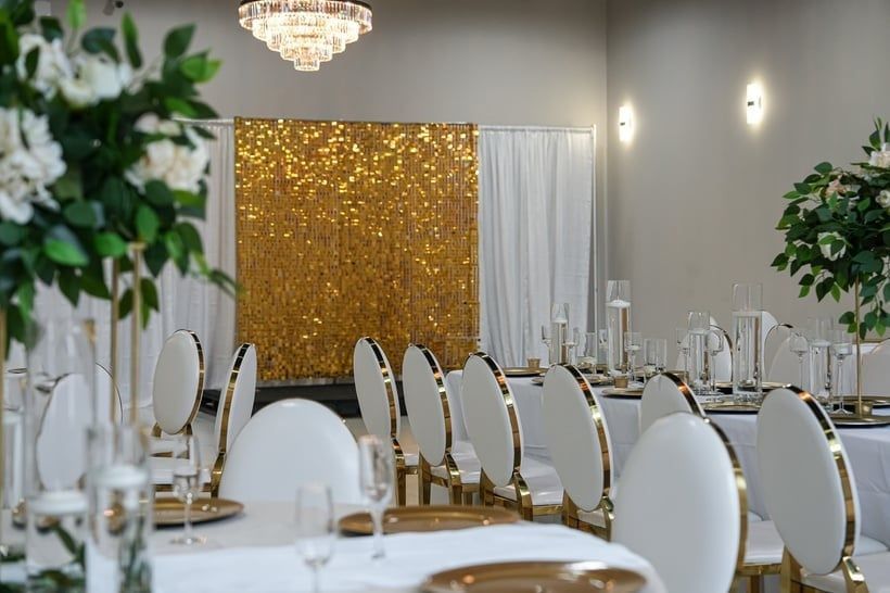 Elegant event space with gold and white decor: chairs, tables, backdrop, and floral arrangements.