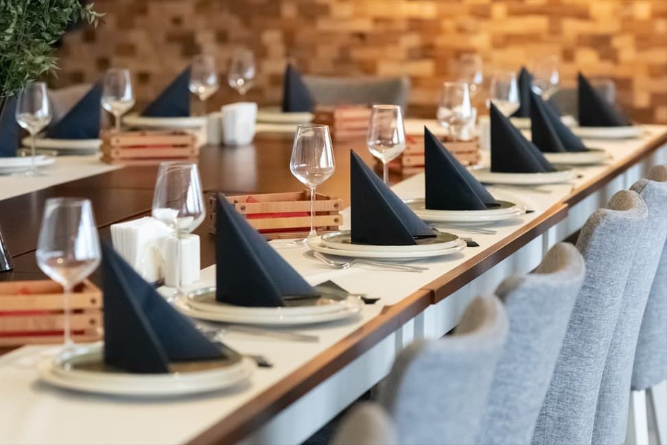 Long wooden table, plates, black napkins, wine glasses, and gray chairs are arranged.