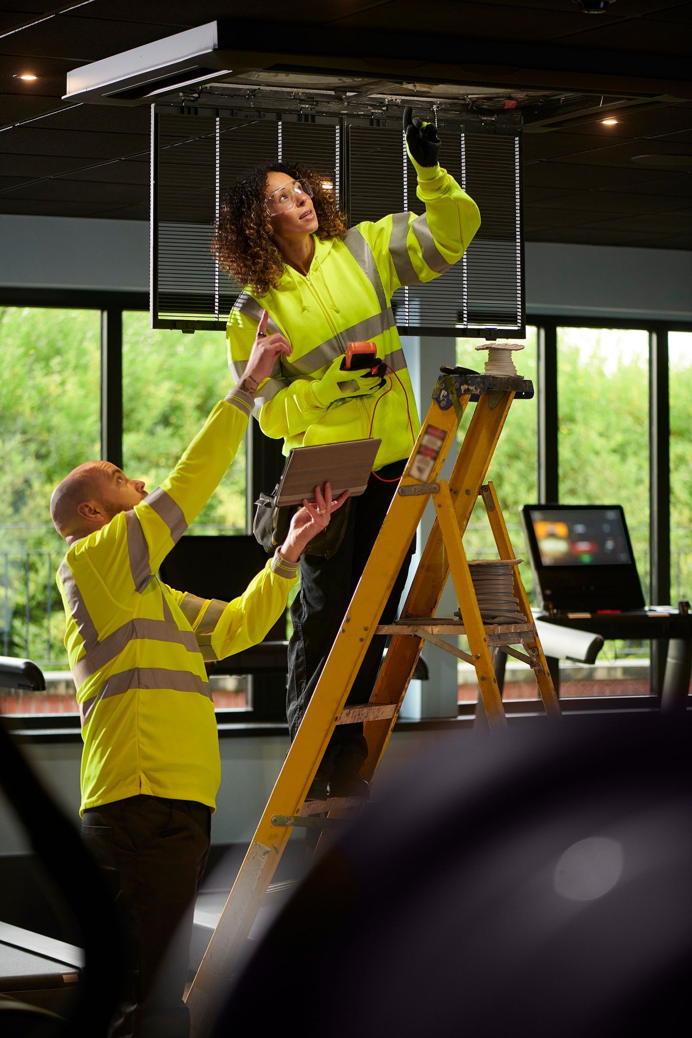 Two workers in safety vests, one on a ladder, adjusting overhead lighting Two workers in safety vests, one on a ladder, adjusting overhead lighting