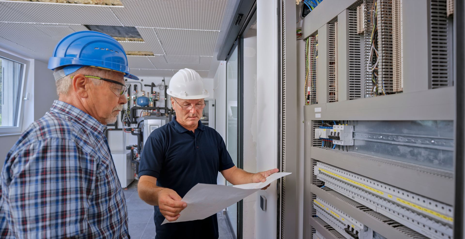 Two people in hard hats examine an electrical panel, one holding a blueprint Two people in hard hats examine an electrical panel, one holding a blueprint