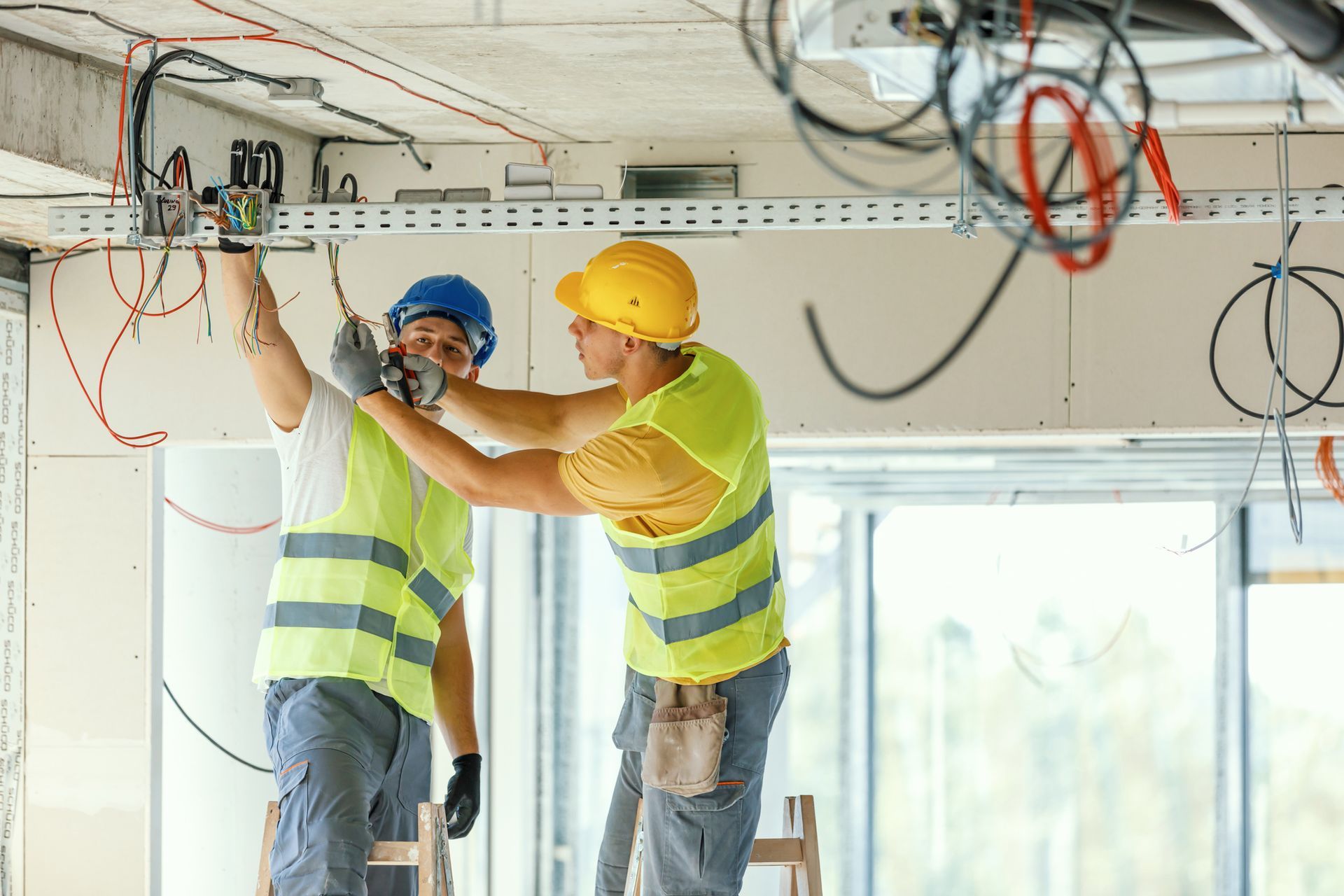 Two electricians in safety gear are wiring electrical cables in a construction site