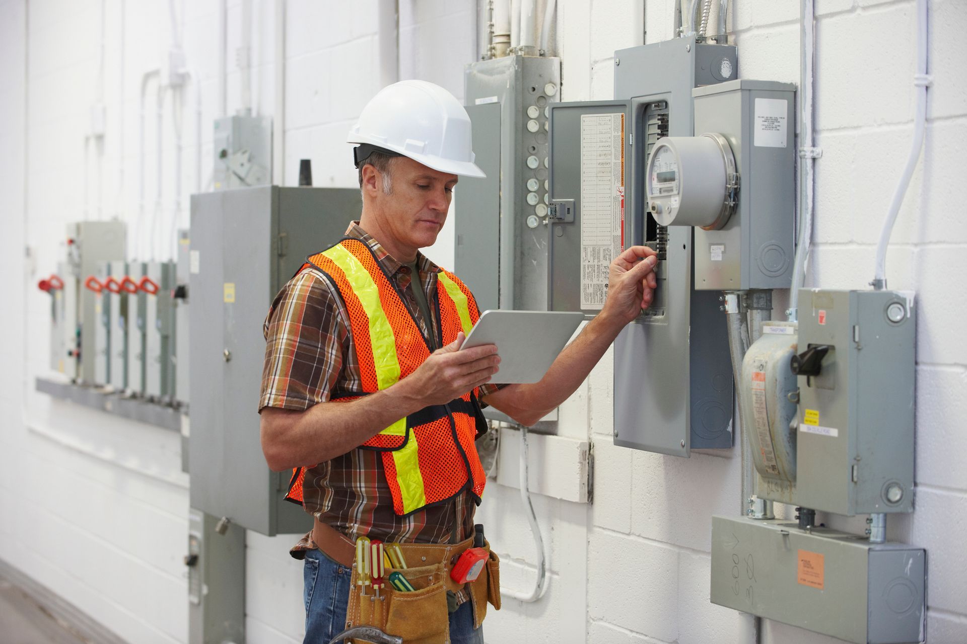 An electrician in a hard hat and vest inspects the electrical panel, using a tablet An electrician in a hard hat and vest inspects the electrical panel, using a tablet