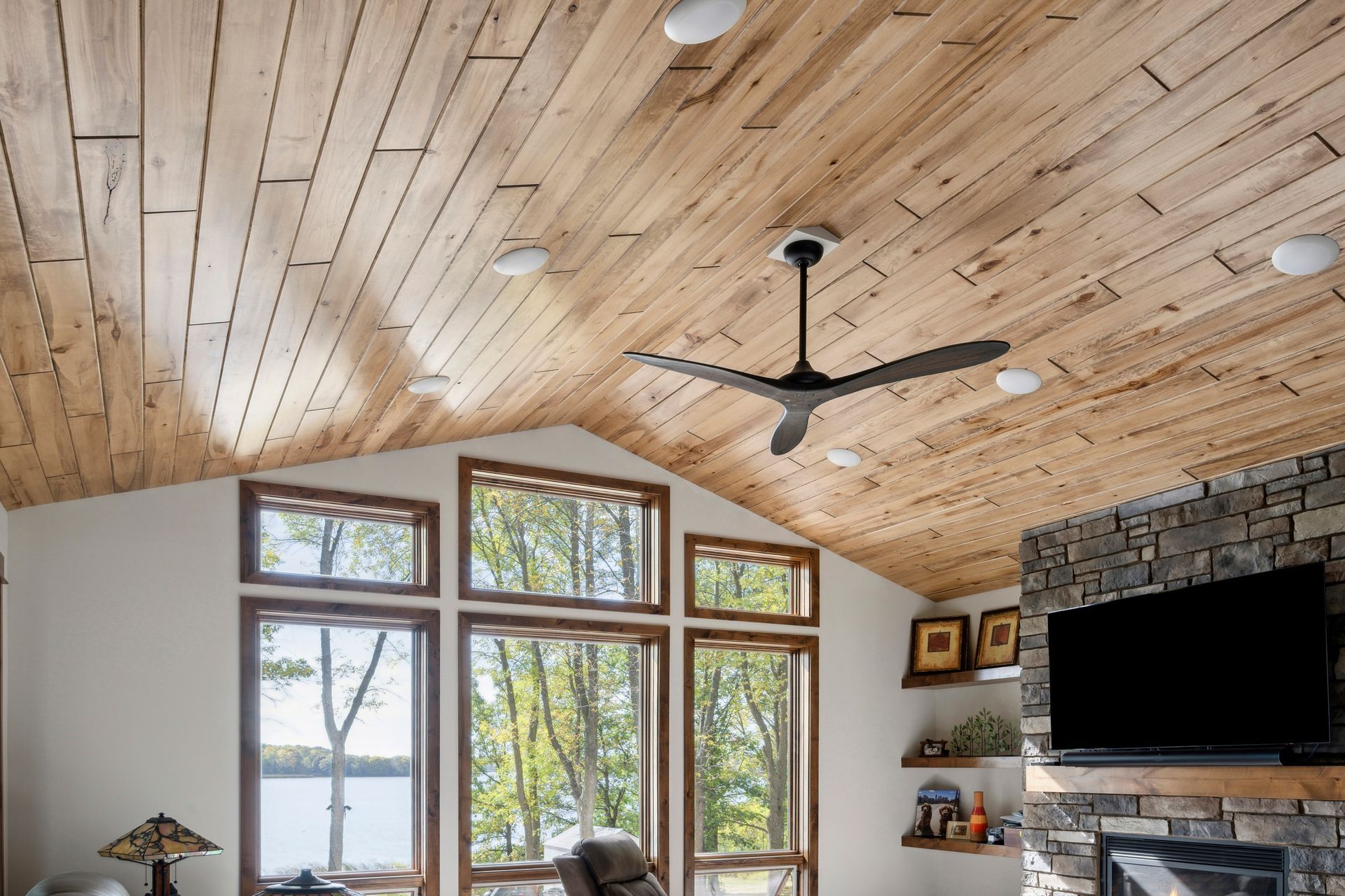 Living room with vaulted wood ceiling, large windows, stone fireplace, and wall-mounted TV