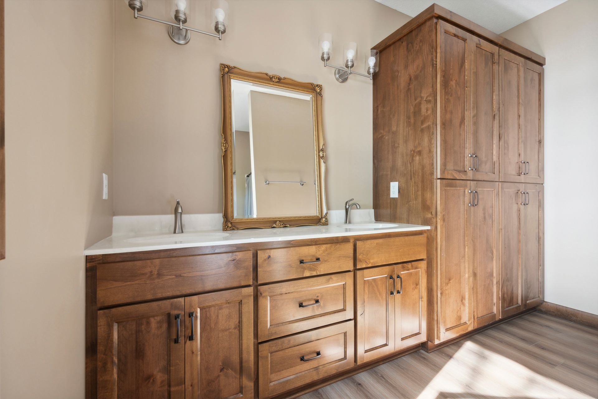 Wood vanity with double sinks, mirrored cabinet, wall sconces, and a tall wood storage cabinet in a bathroom.