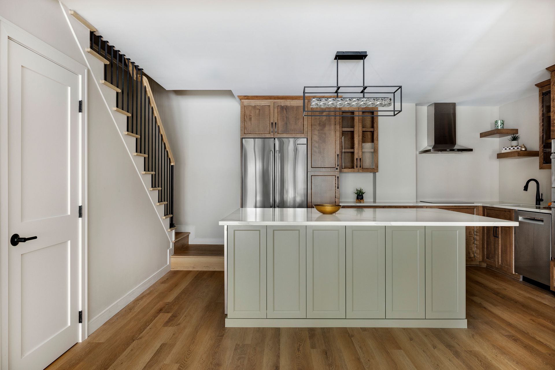 Modern kitchen with white island, wood floors, staircase, and stainless appliances