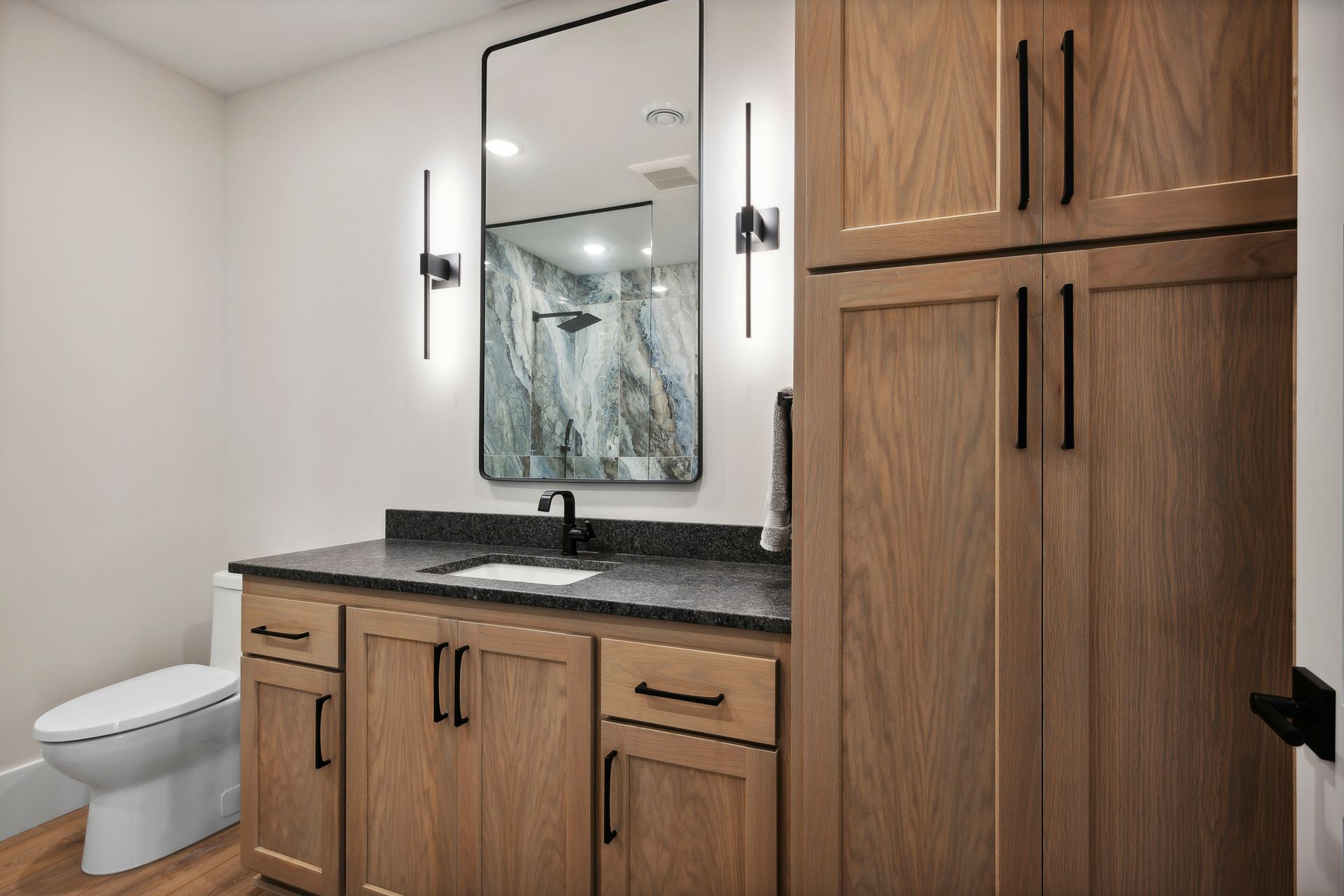 Modern bathroom vanity with wood cabinets, black countertop, mirror, sink, and toilet