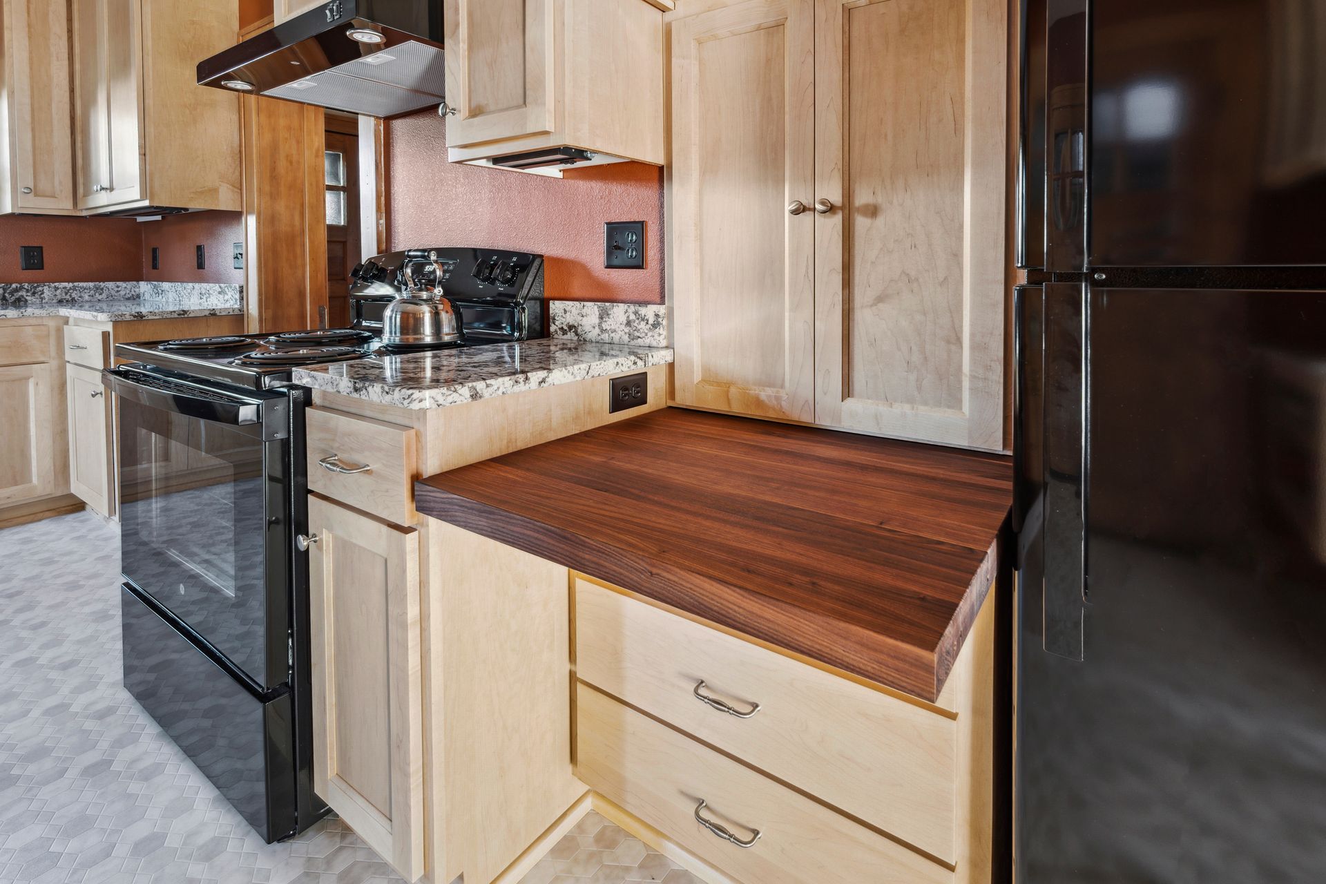 Kitchen with light wood cabinets, black appliances, and a butcher-block countertop beside a stove.