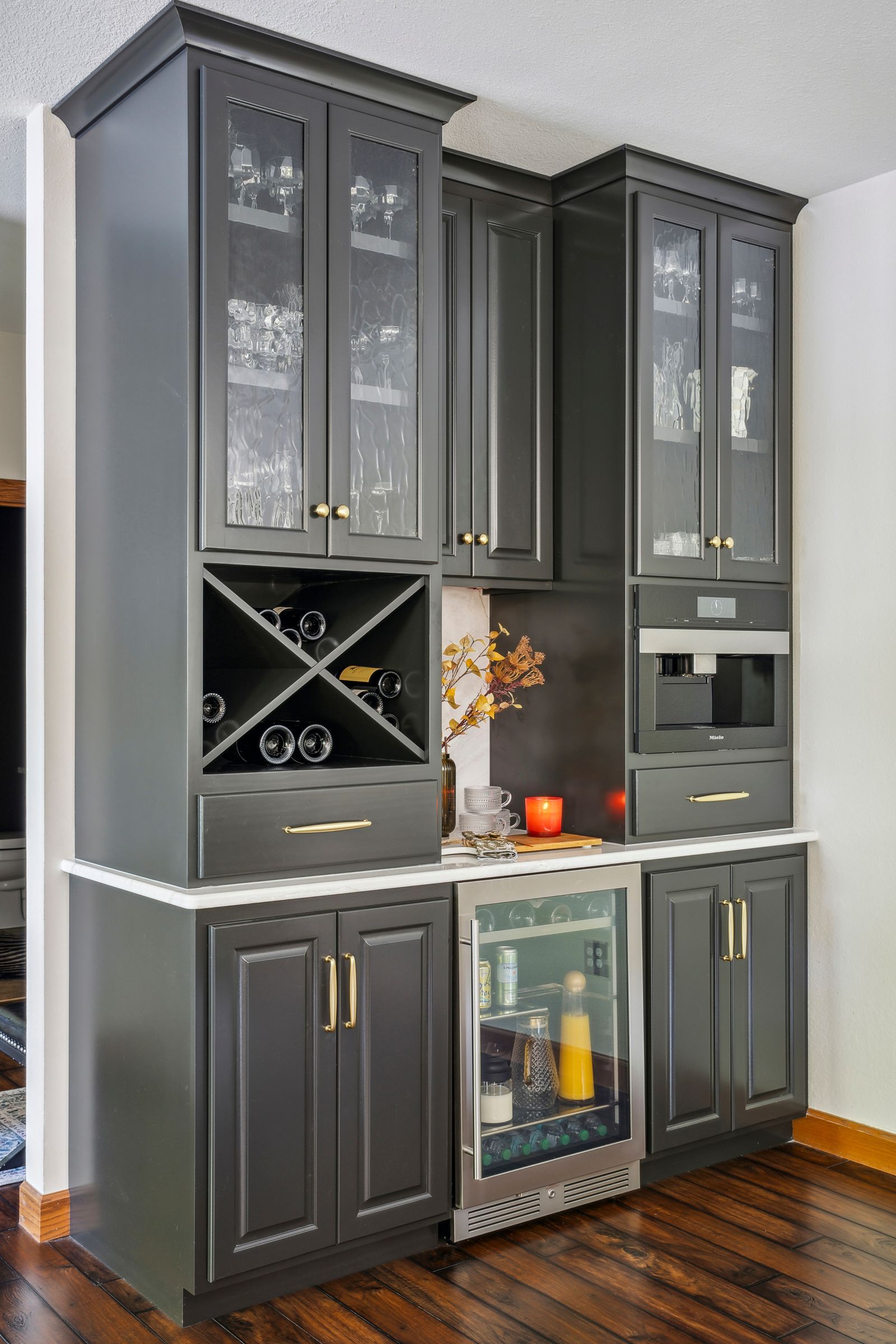 Gray built-in kitchen cabinet with glass doors, wine rack, and wine fridge against a white wall.