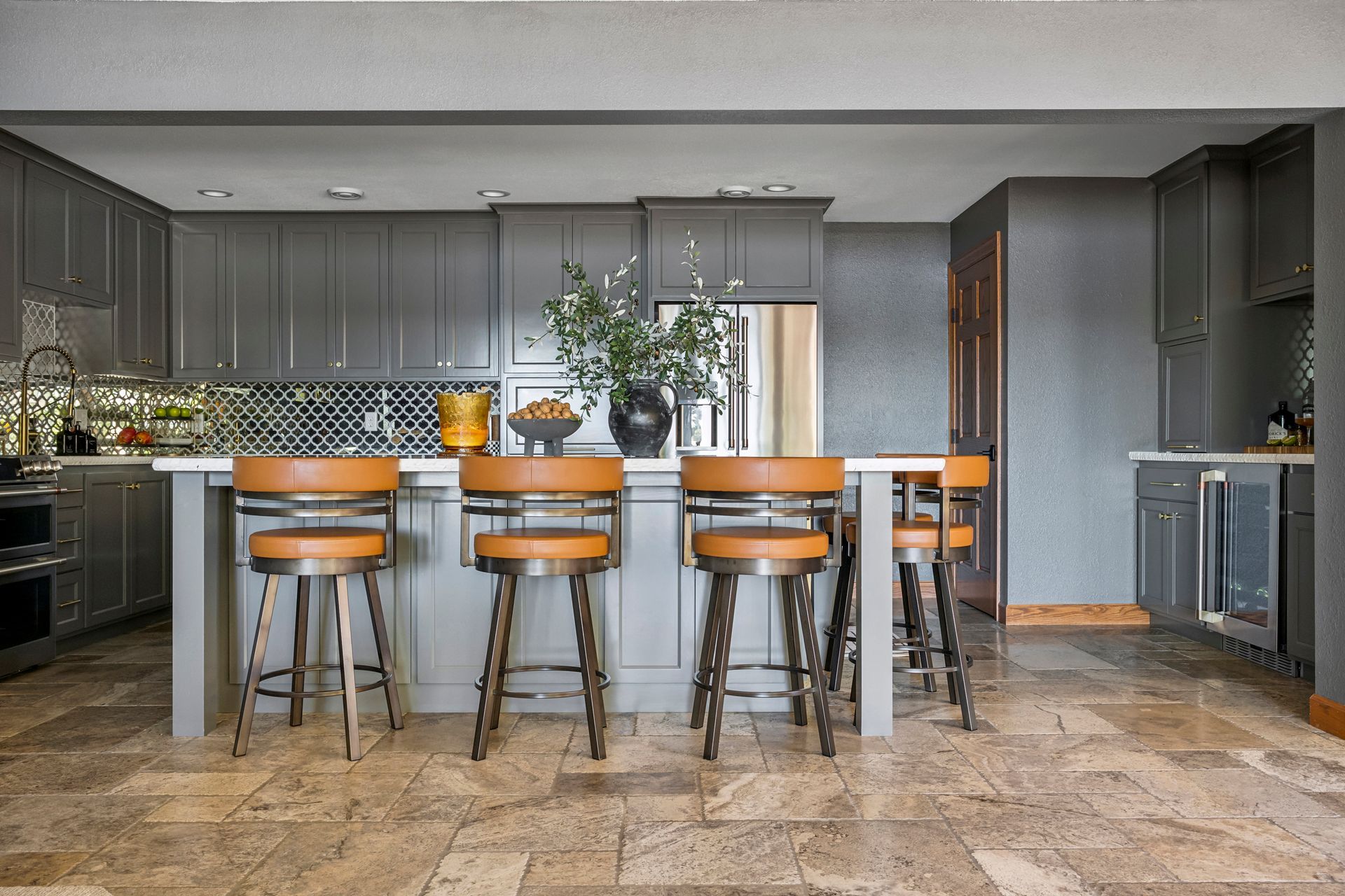 Modern kitchen with dark cabinets, stone floor, and a long island with orange stools and a vase centerpiece
