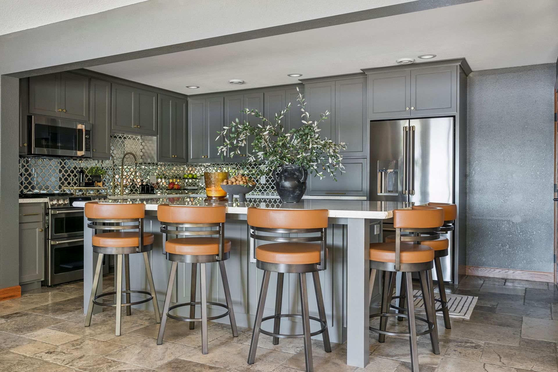 Modern kitchen with a large island, orange bar stools, gray cabinets, and a stainless steel refrigerator.