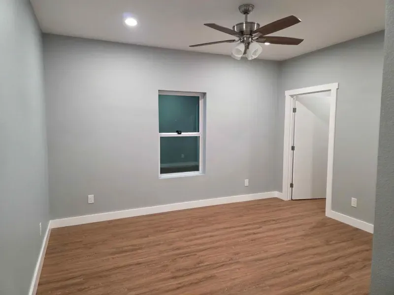 Empty gray bedroom with wood flooring, ceiling fan, window, and an open door