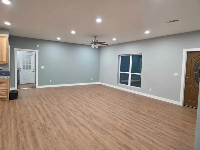 Empty living room with gray walls, wood flooring, recessed lights, ceiling fan, window, and front door.