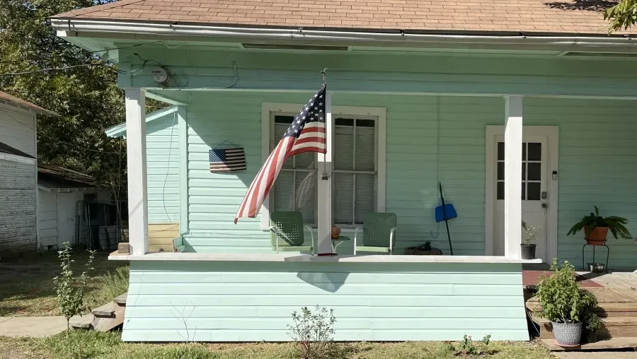 Mint-green house porch with white columns, an American flag, and potted plants in front.