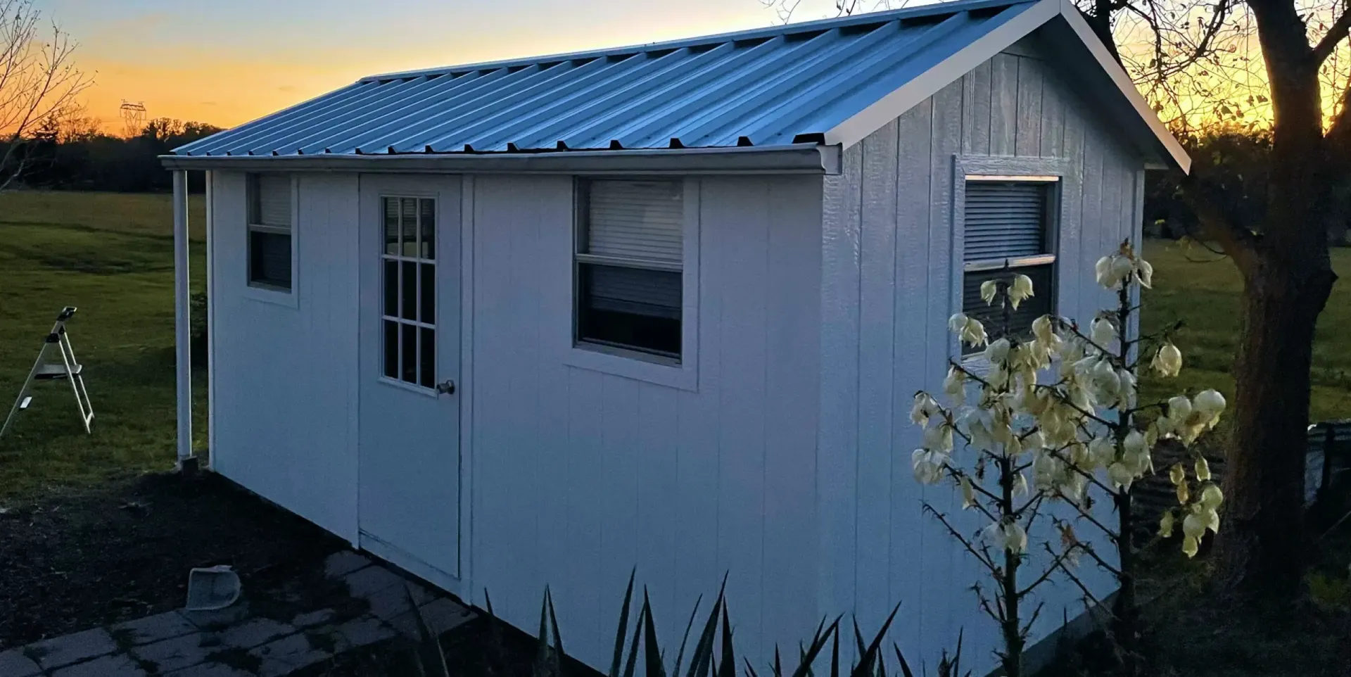 Small white shed with a metal roof at sunset in a grassy yard