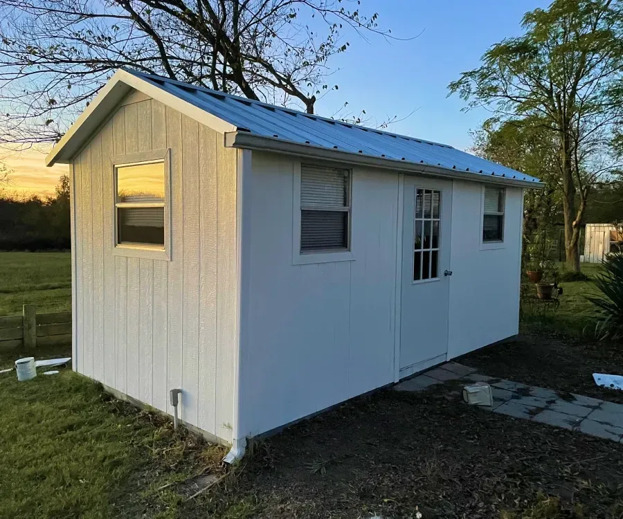 Small white shed with a blue metal roof and windows, standing on a grassy yard at sunset.
