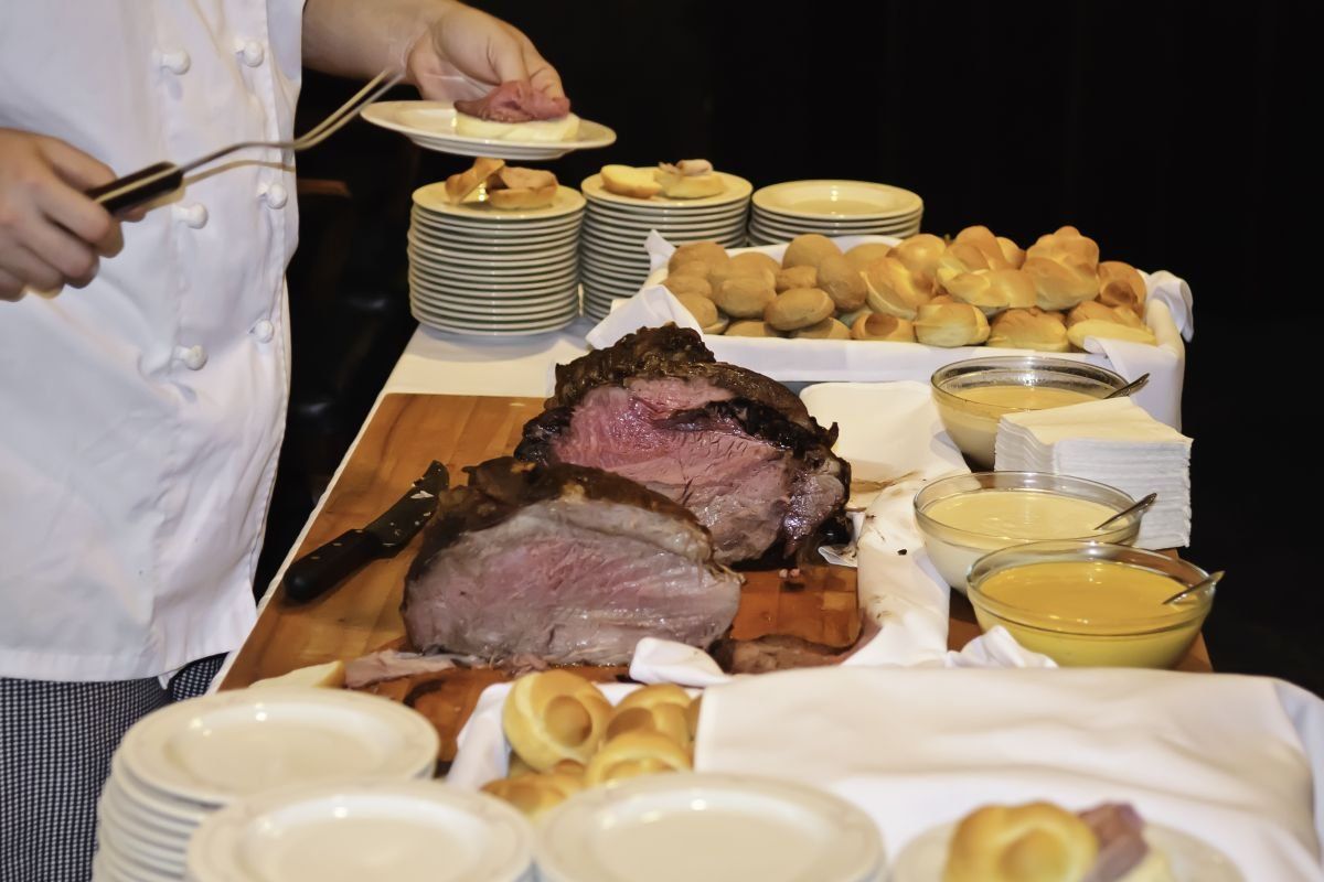 Carved roast beef at a buffet with chef serving potatoes and gravy, plates stacked nearby