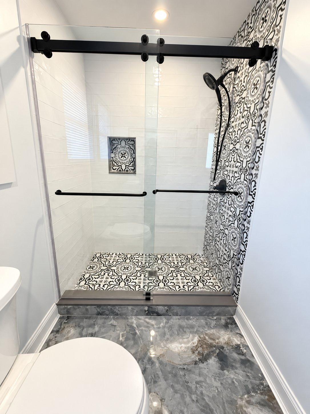 Small tiled shower with black grab bars, patterned bench, and shower curtain in a narrow bathroom.