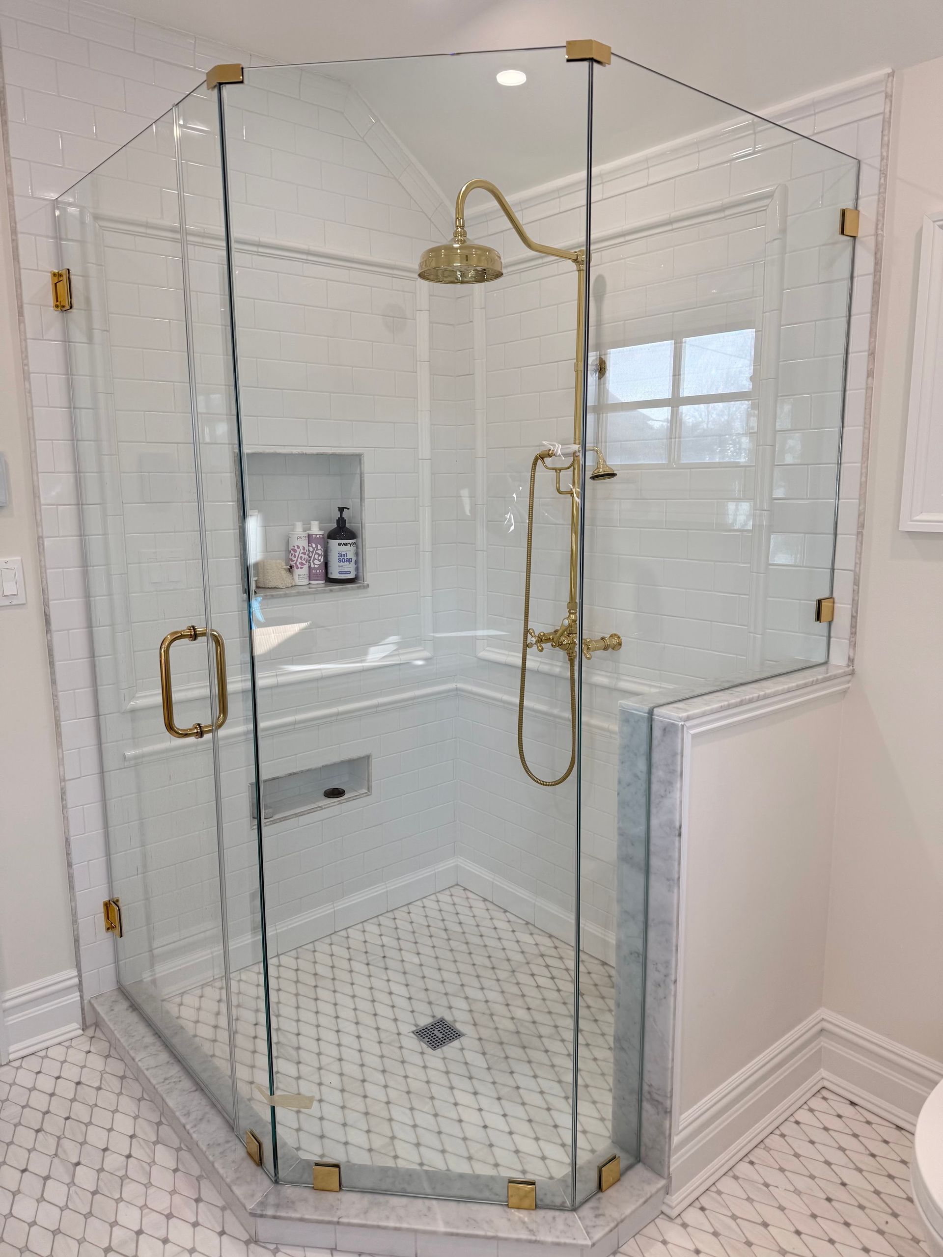 Glass-enclosed corner shower with brass fixtures and white mosaic tile floor