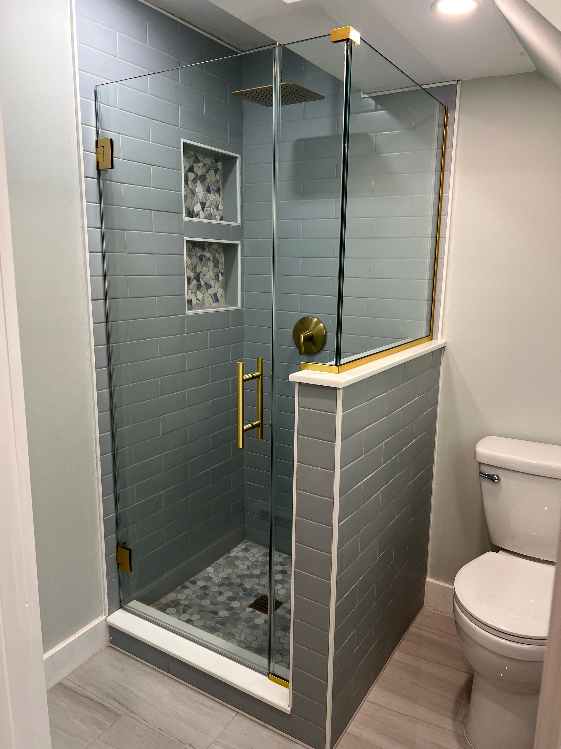 Glass shower enclosure in a bathroom with gray tile and brass hardware, next to a toilet.