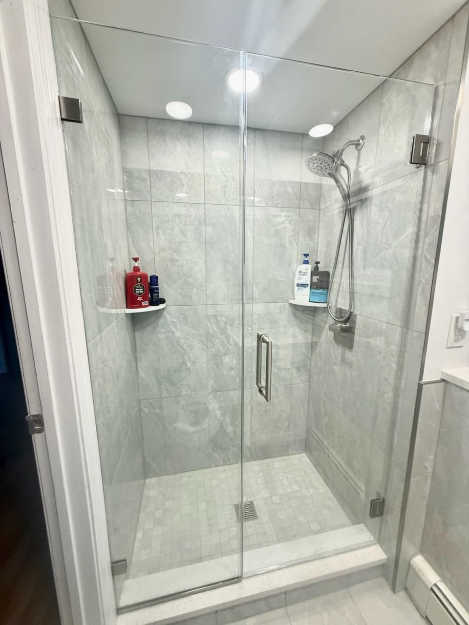Glass-enclosed shower with gray tile, chrome fixtures, and corner shelves holding toiletries