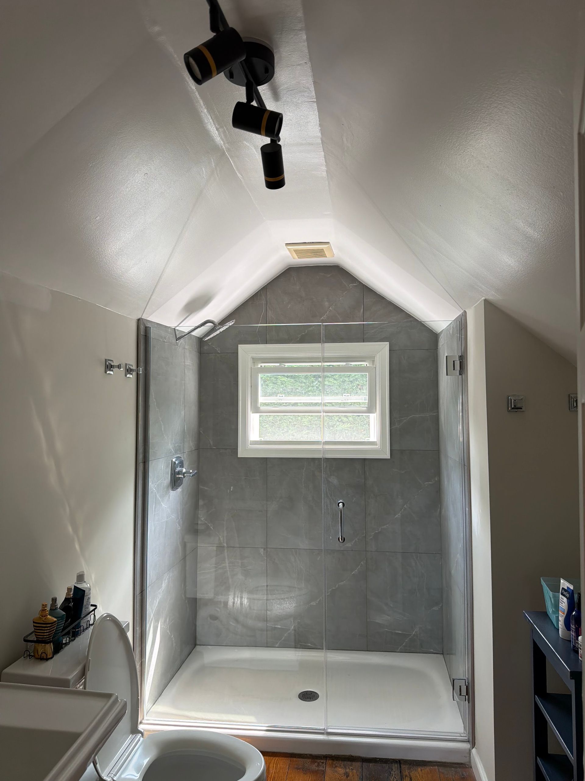 Modern bathroom shower with gray tile, white sloped ceiling, and a small window above the tub.