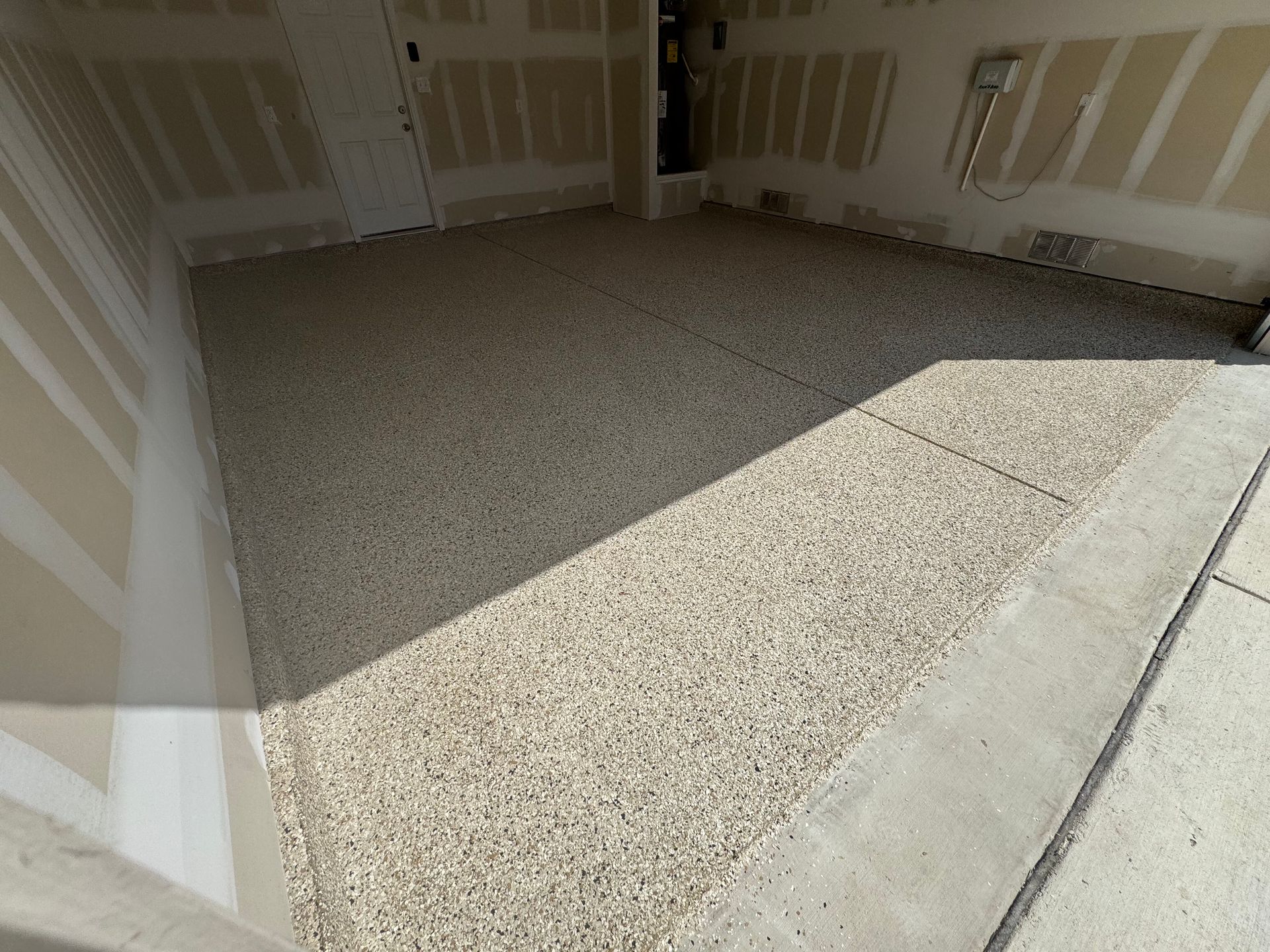 Empty unfinished garage with drywall walls and a gravel floor, lit by sunlight.