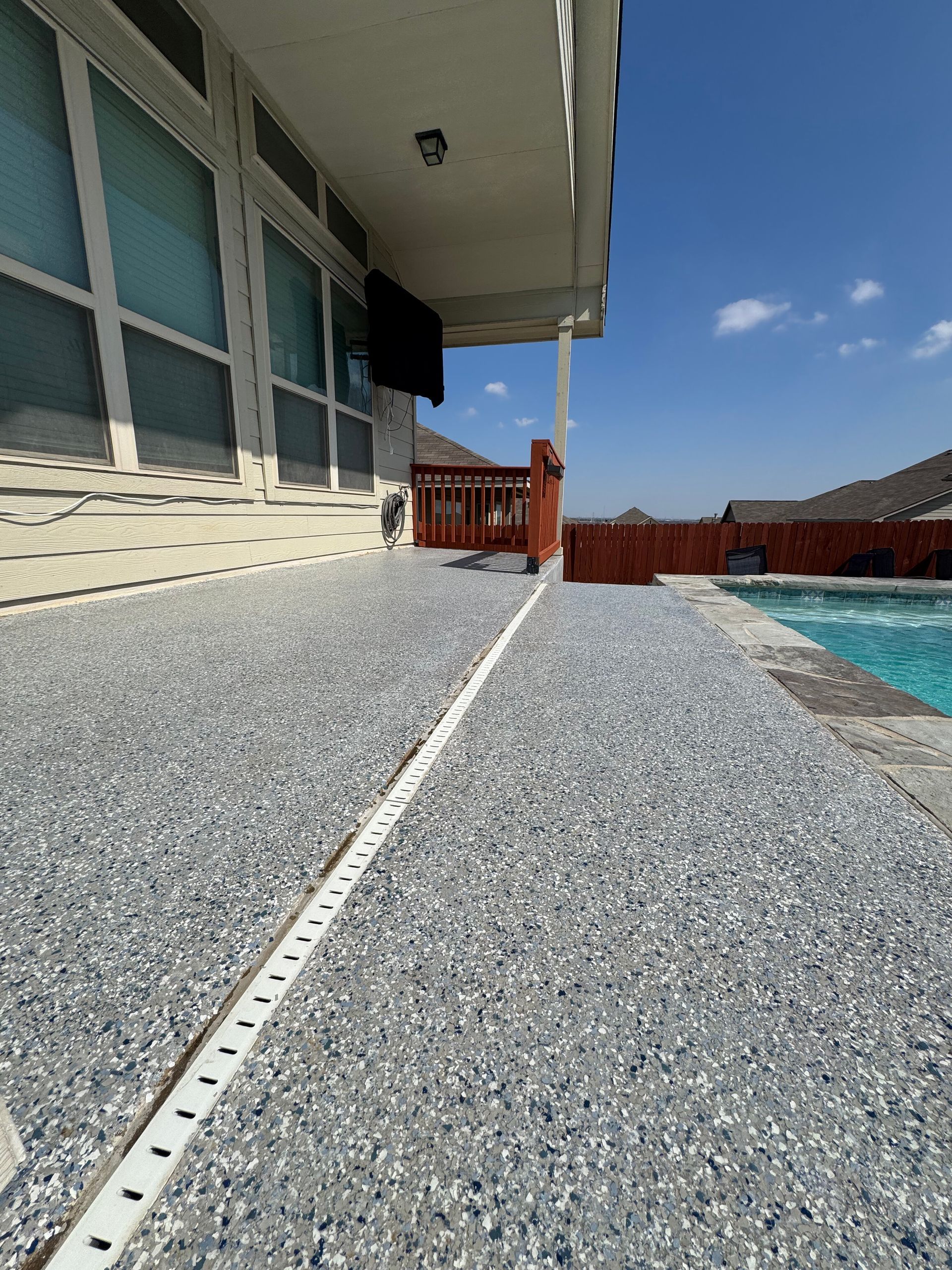 Poolside patio with textured concrete walkway, house windows, and blue pool under a clear sky