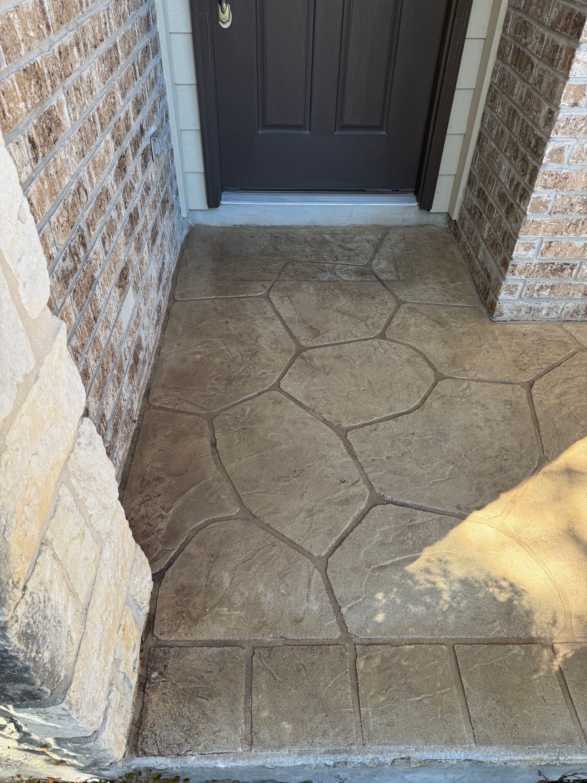 Stone front porch with dark brown door and textured walls, lit by sunlight.
