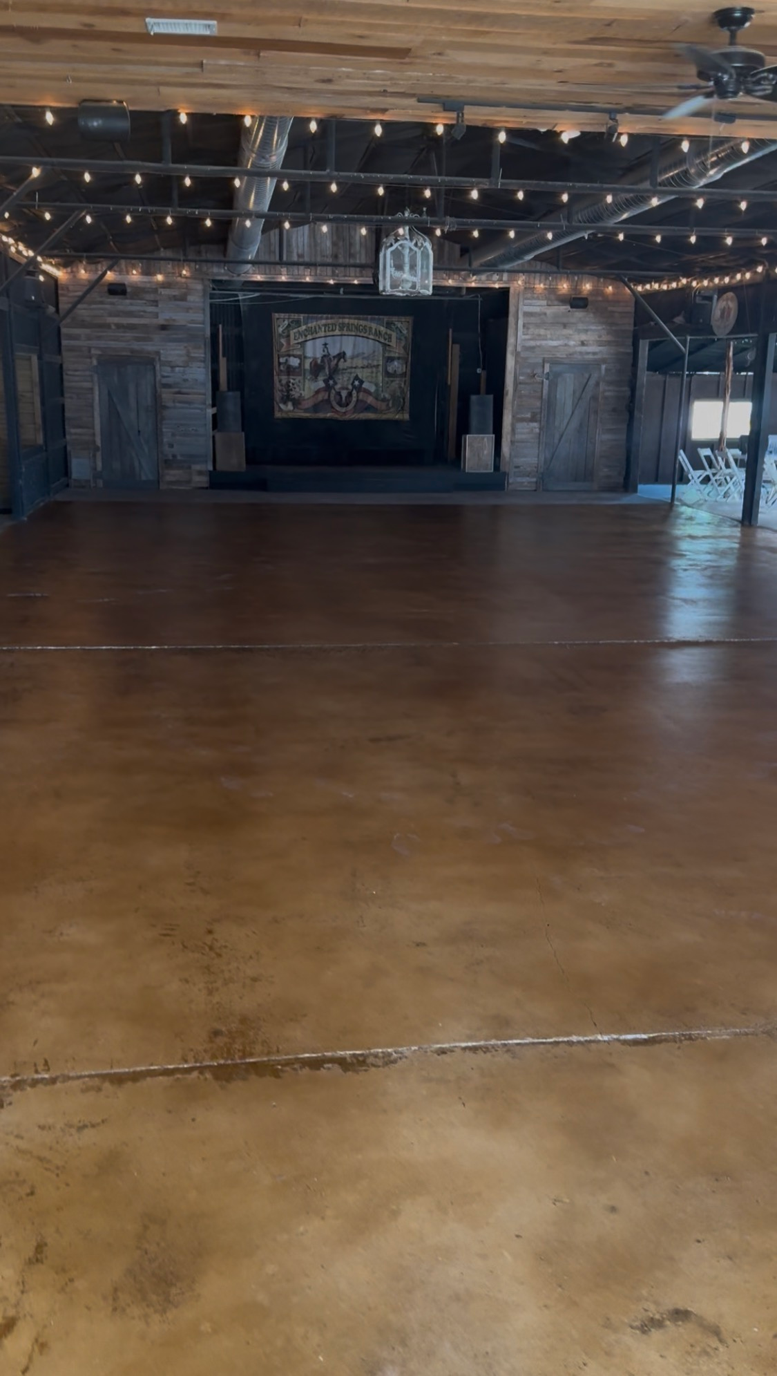 Empty rustic event hall with wooden floor, string lights, and stage area at the back