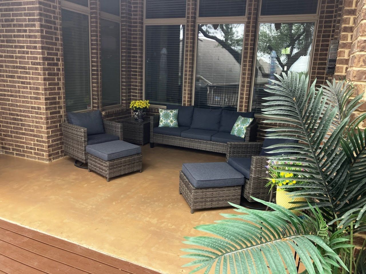 Brick patio with dark gray outdoor sofa, ottoman, and potted plants beside large windows