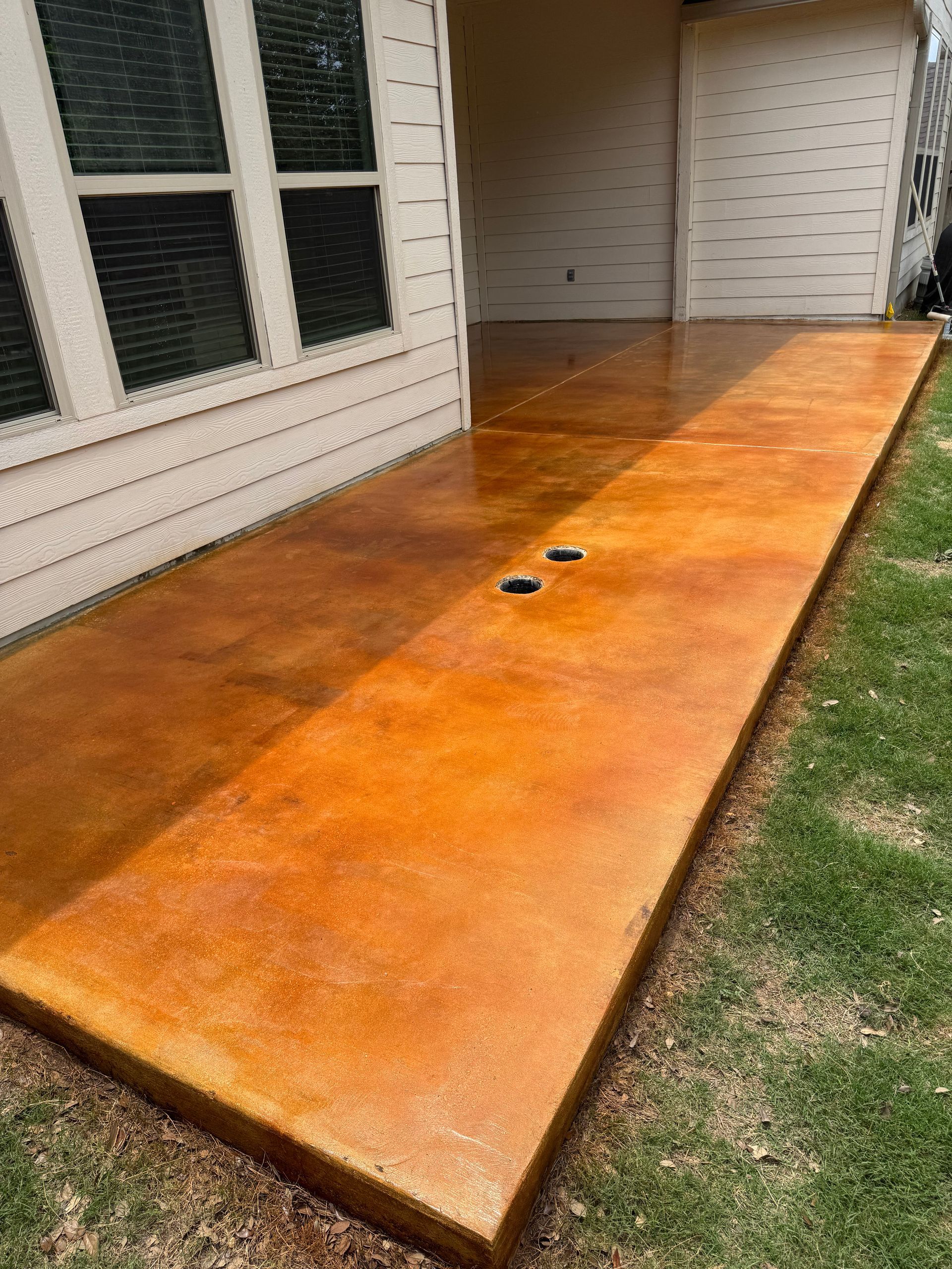 Orange-stained concrete patio beside a house with windows, bordered by grass and a garage opening