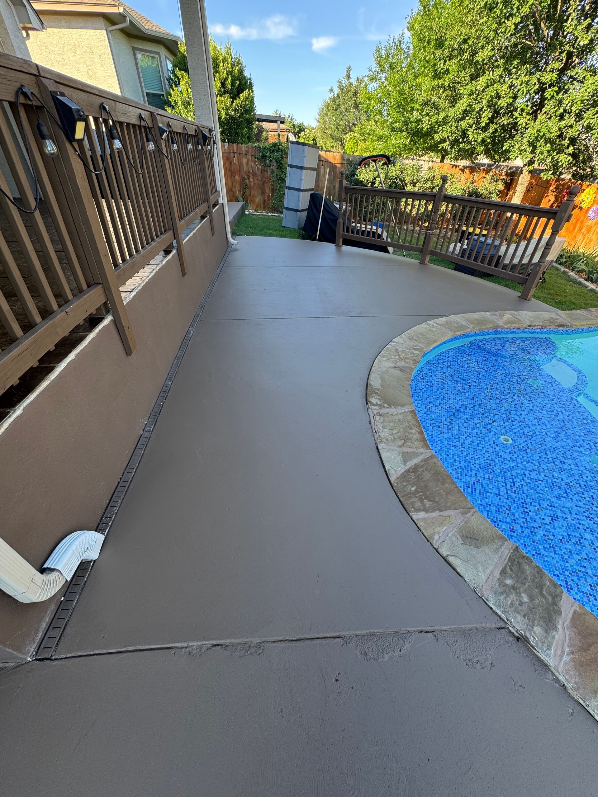 Concrete pool deck beside a blue swimming pool, bordered by a wooden fence and trees.