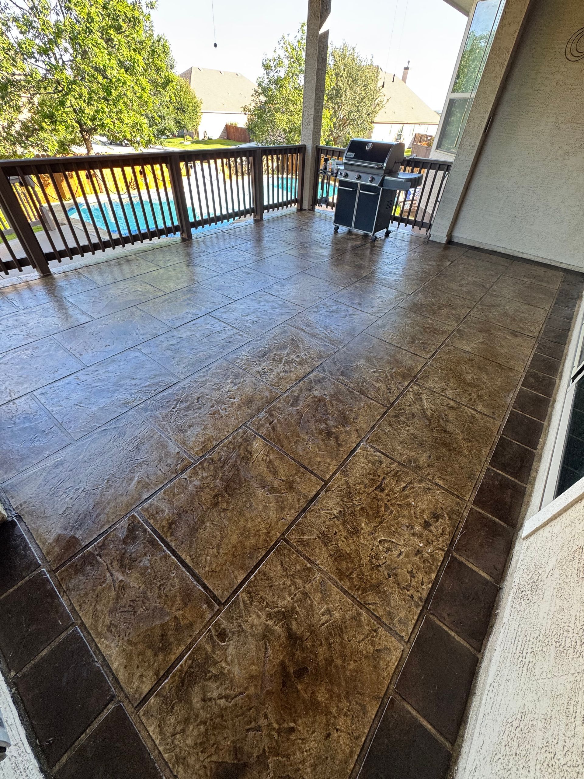 Covered patio with wet stamped concrete floor and black railing overlooking a pool area