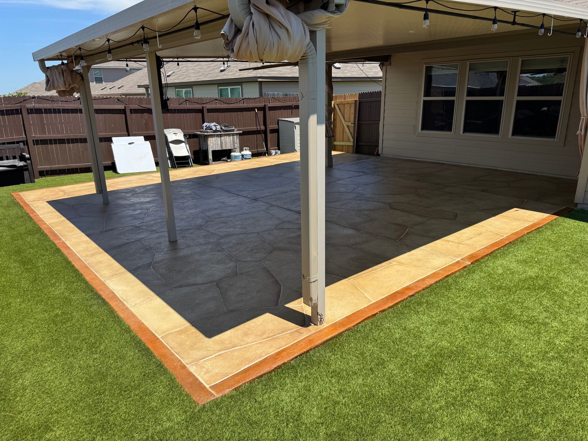 Covered patio with dark floor and tan trim beside a house, viewed from the yard