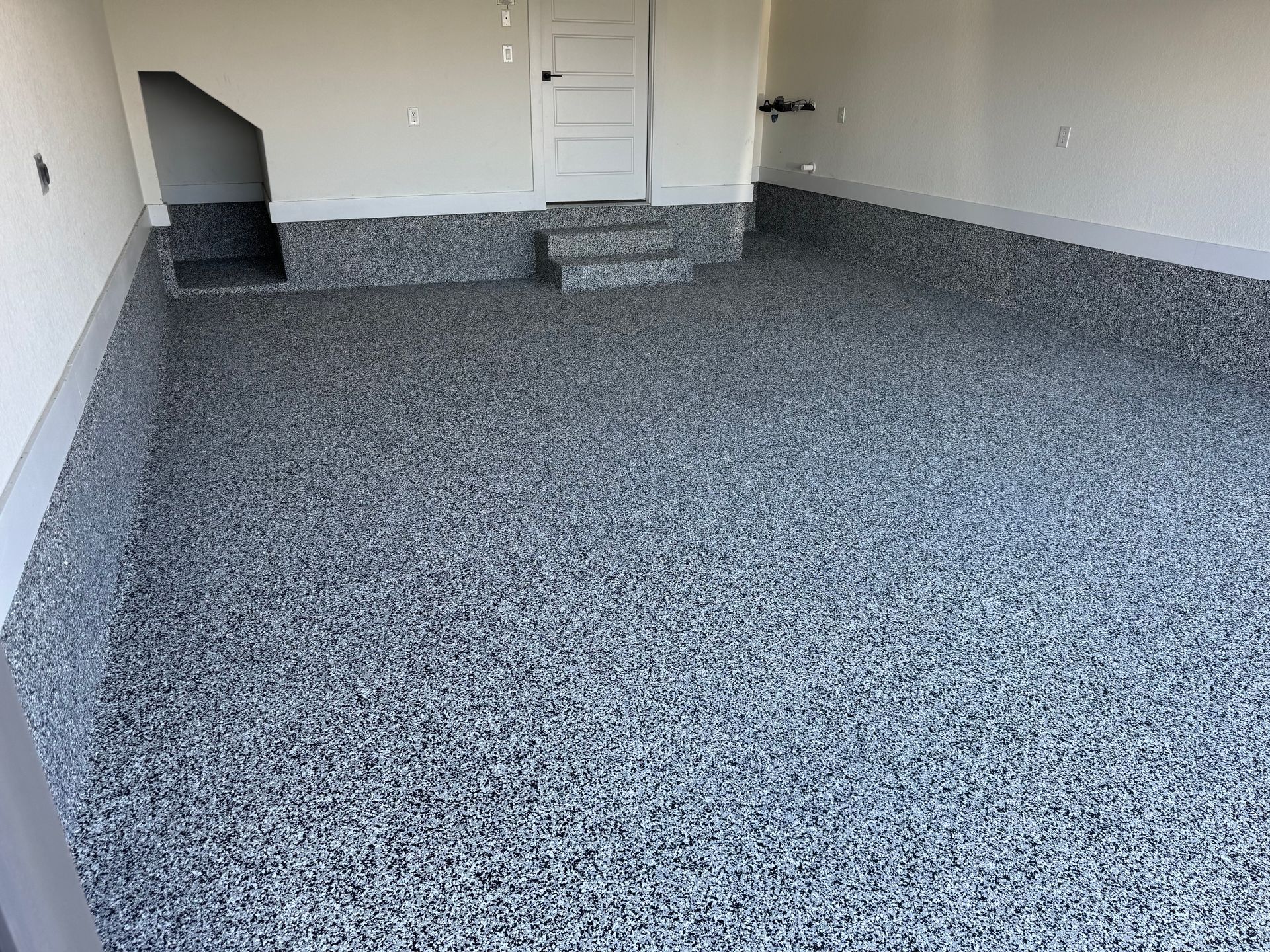 Empty carpeted living room with gray speckled floor and white walls, leading to a small stairway and door.