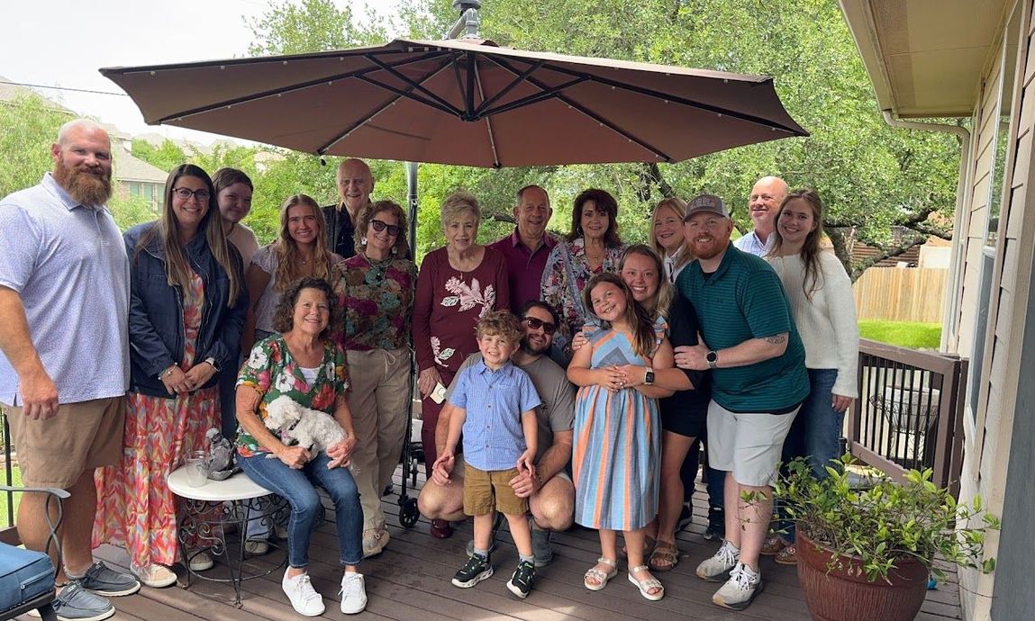 Group of people posing together under a patio umbrella on a porch, smiling at the camera.