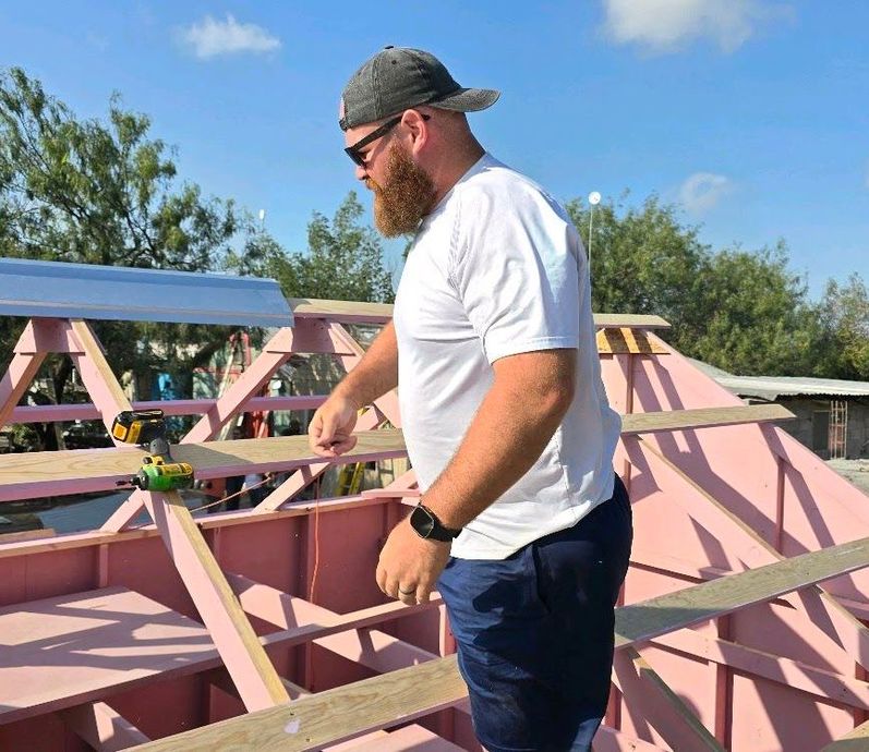 Man in white shirt and cap inspecting wooden roof framing on a pink structure under a blue sky