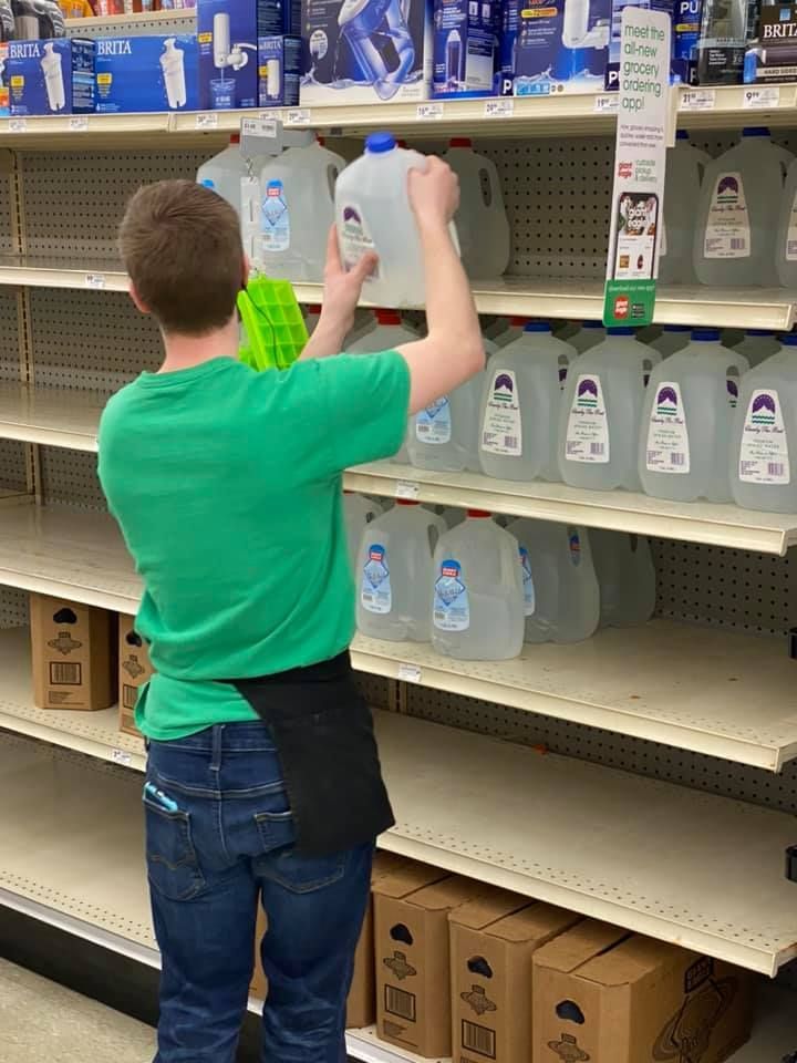 A person in a green shirt stocks water jugs on a mostly empty store shelf. Boxes are on the bottom shelf.