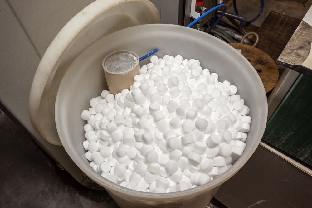 White salt pellets fill an open, cylindrical container. A small scoop and cardboard cup sit inside.