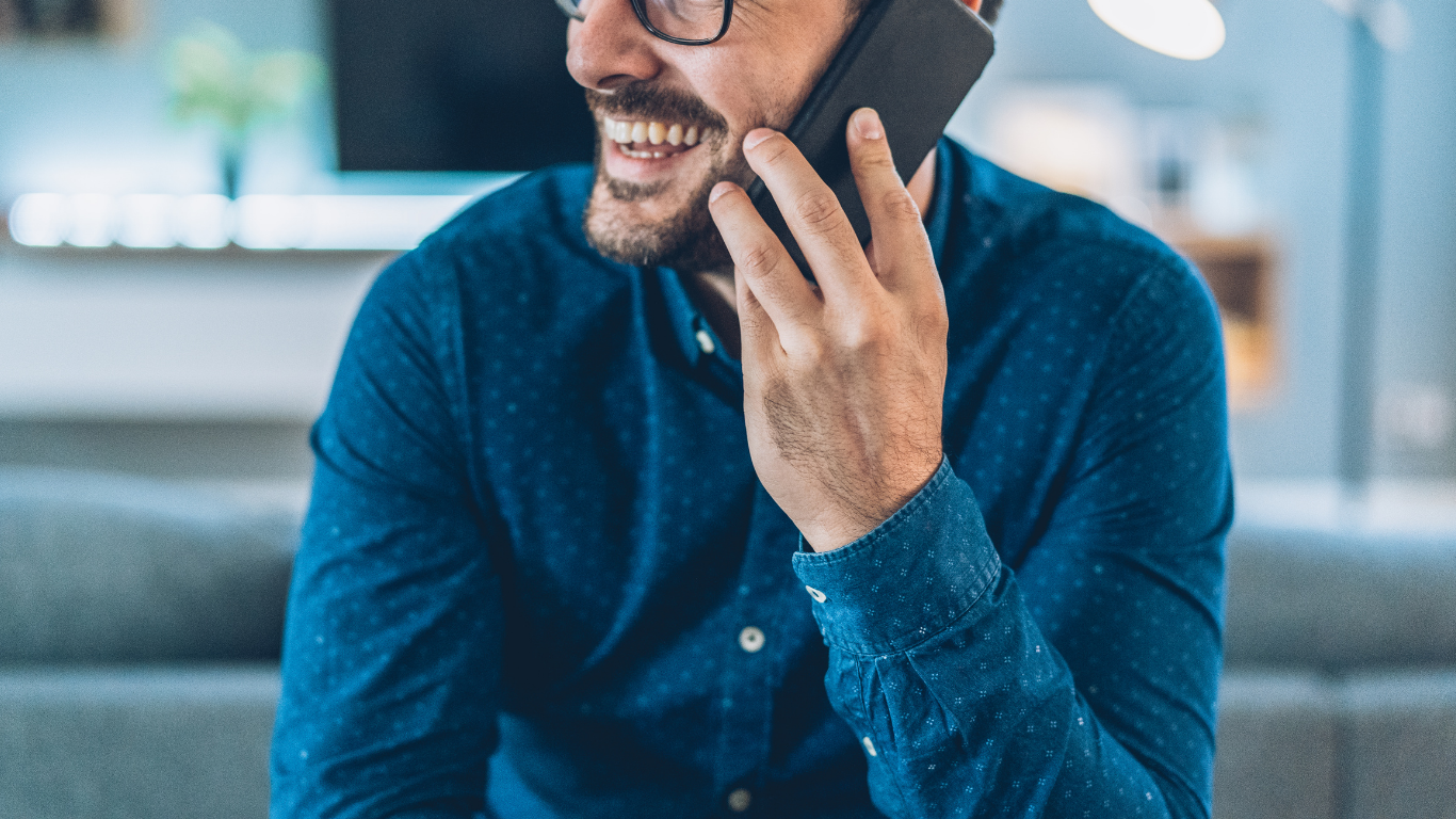 Man in blue shirt smiling while talking on a smartphone indoors
