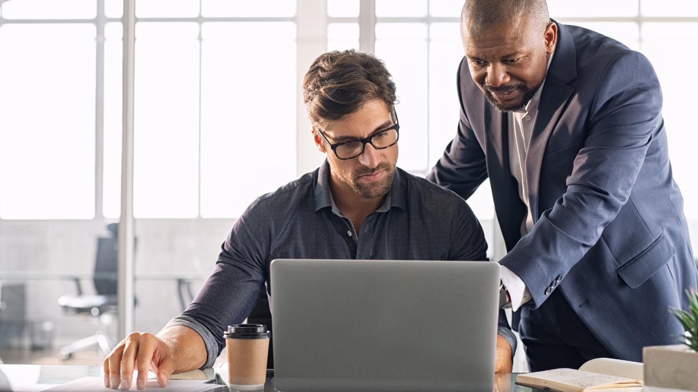 Two coworkers reviewing a laptop in a bright office, one standing and pointing over the seated man's shoulder.