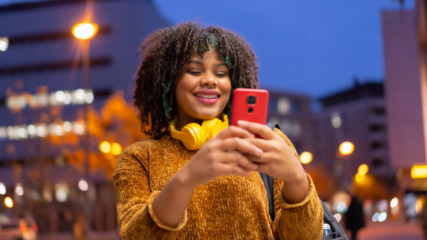 Smiling person in a brown sweater takes a selfie with a red phone at night, wearing yellow headphones.