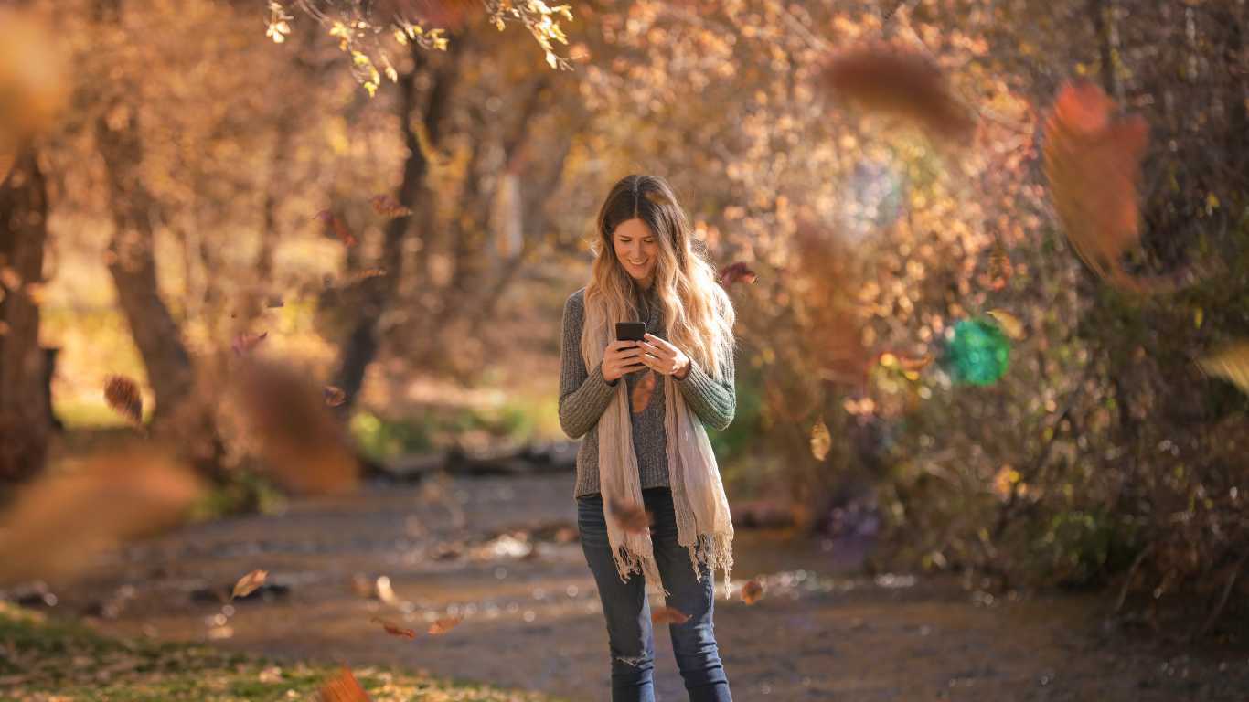 Person standing on a tree-lined path, looking at a phone amid warm autumn foliage.