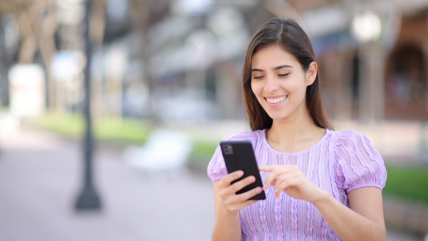 Smiling woman in a lavender dress looking at her phone outdoors on a blurred city street.