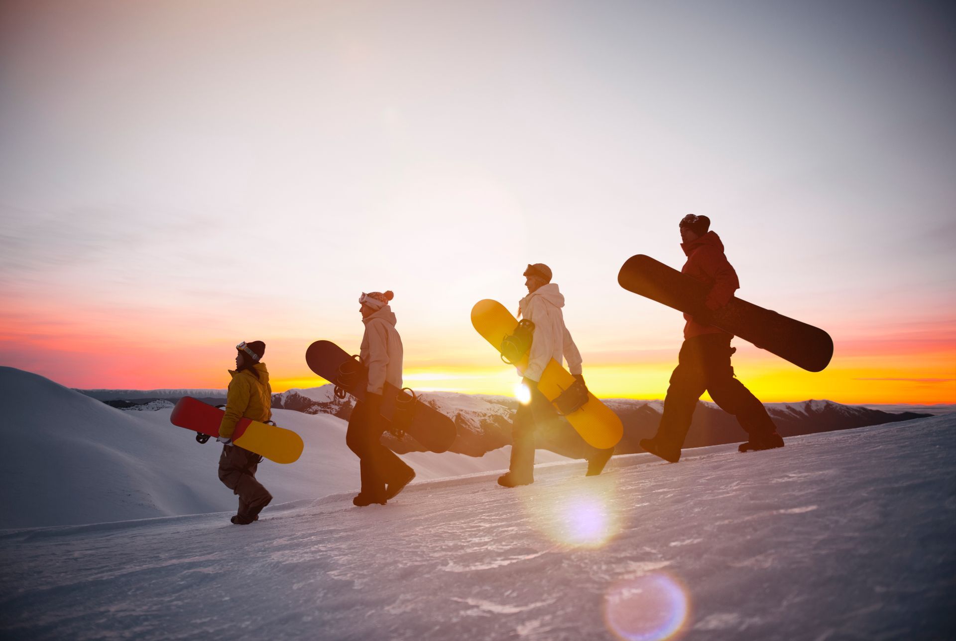A group of snowboarders walking on top of a mountain at sunset. They are holding their snowboards.