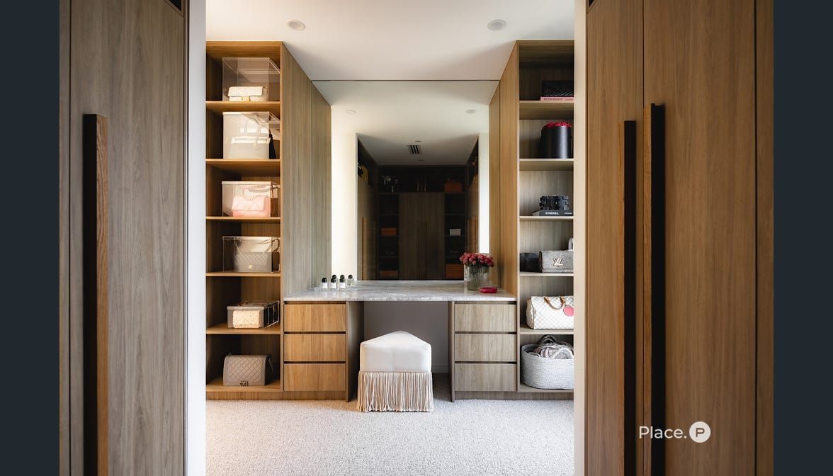 A walk-in closet featuring light wood shelving, a central vanity with a stool, and a large mirror framed by cabinet doors. — Foreshore Kitchens in Coolum Beach, QLD