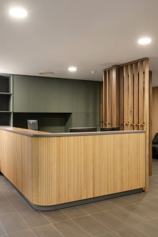 A modern reception desk with light wood panelling and curved edges, set against a matte olive-green wall and wood screen. — Foreshore Kitchens in Coolum Beach, QLD
