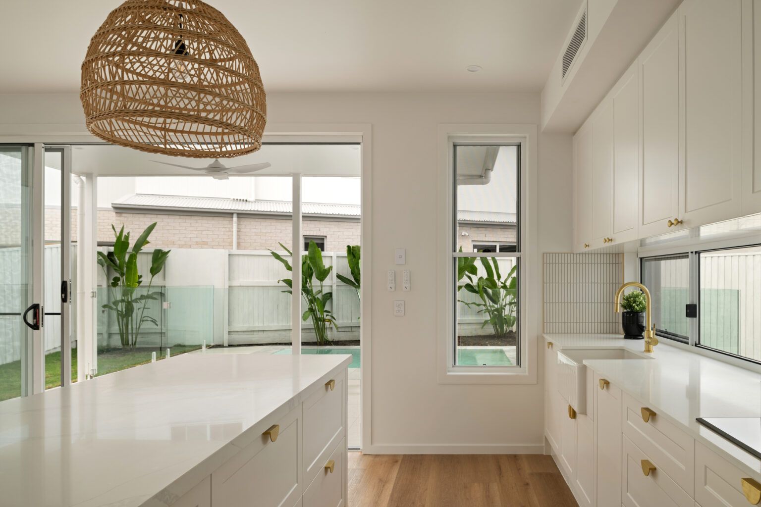 A bright, modern white kitchen with quartz countertops, gold hardware, and a woven pendant light overlooking a pool. — Foreshore Kitchens in Coolum Beach, QLD