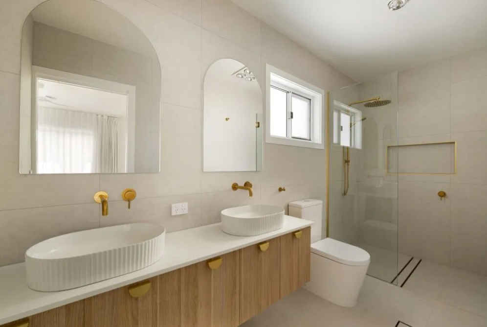 Bathroom with double vanity, arched mirrors, gold tapware, basins, toilet and glass shower